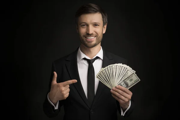 Usmívající se podnikatel ukazující prst na peníze v ruce, černé pozadí — Stock fotografie