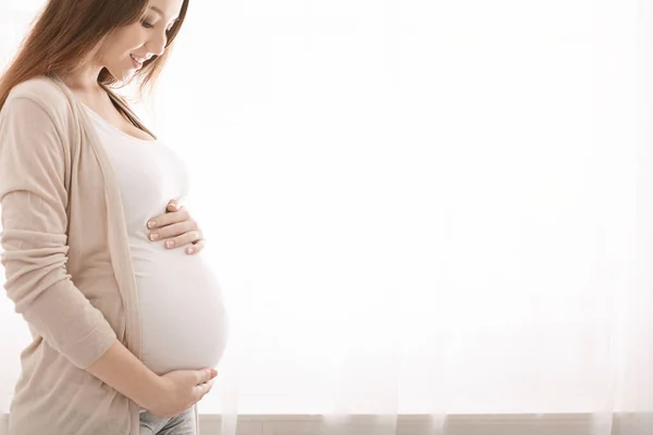Счастливая беременная женщина дотрагивается до живота у окна — стоковое фото