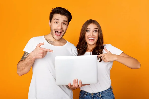 Excited millennial couple pointing on laptop, orange background