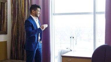 Yakışıklı takım elbiseli adam otelin penceresinin yanında kahve içiyor.