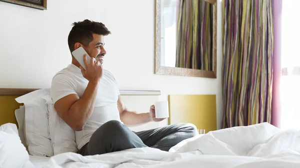 Mladý muž sedí na posteli, telefonuje, pije čaj — Stock fotografie