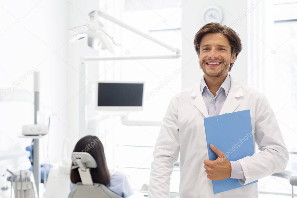 Delighted dentist smiling and holding folder in dental cabinet