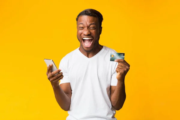 Afro Guy segurando telefone e cartão de crédito gritando, fundo amarelo — Fotografia de Stock