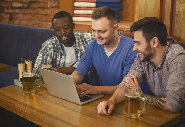 Three young men drinking beer, using lapop in pub together — Stock Photo, Image