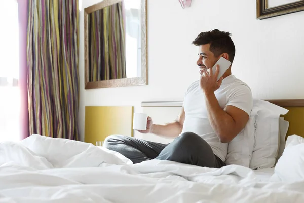 Mladý muž sedí v posteli, mluví na smartphonu, pije kávu — Stock fotografie