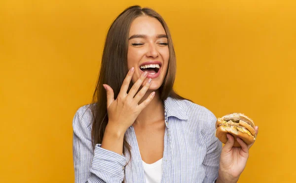 Burger yiyen ve gülen genç bir kadın Sarı Arkaplanda dikiliyor — Stok fotoğraf