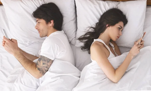 Millennial casal em disputa, deitado na cama de volta para trás, usando celulares — Fotografia de Stock