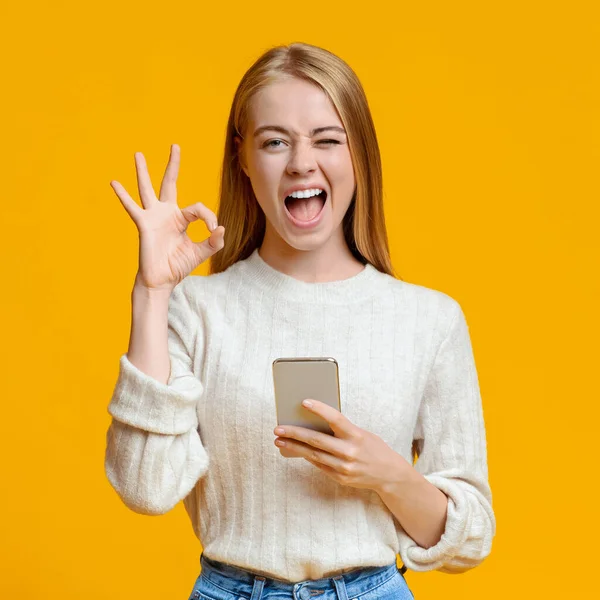 Millennial chica sosteniendo teléfono inteligente, guiño y gesto ok — Foto de Stock