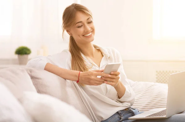 Расслабленная женщина с помощью смартфона сидит с ноутбуком на диване в помещении — стоковое фото