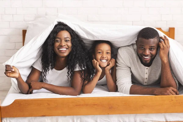 Веселая чёрная семья из троих прячется под одеялом на кровати. — стоковое фото