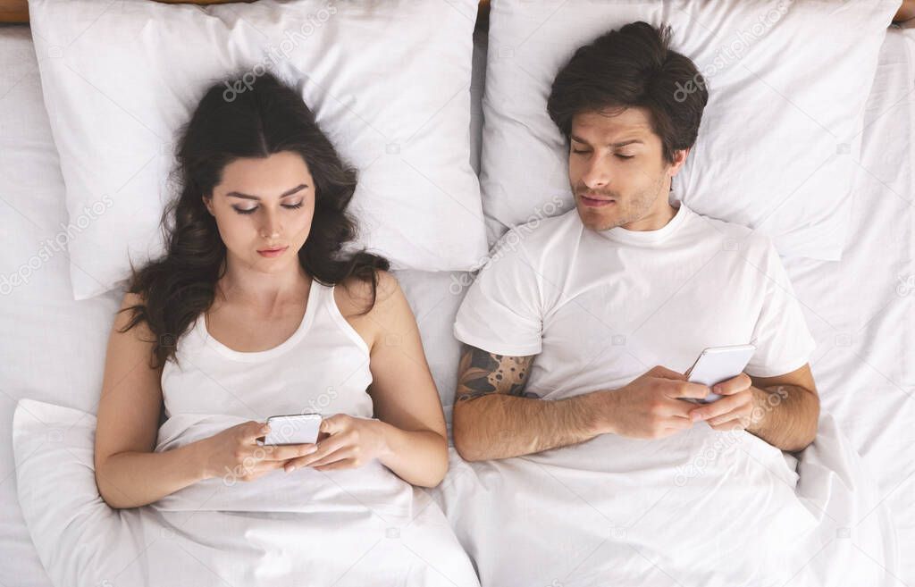 Young couple browsing cellphones in bed, man peeking into wifes phone