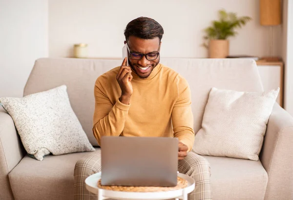 Afro hombre usando el ordenador portátil tener agradable conversación telefónica en casa — Foto de Stock