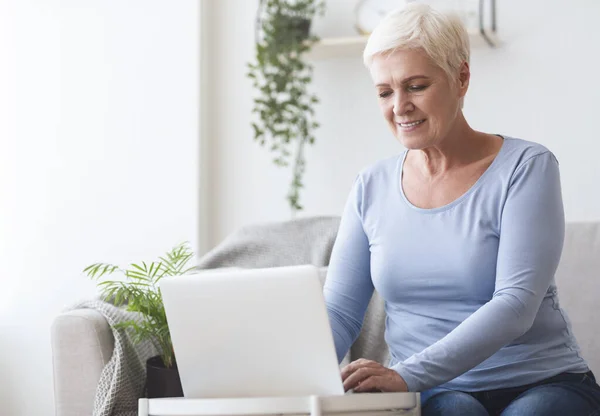 Vrolijke senior dame met behulp van laptop op de bank thuis — Stockfoto