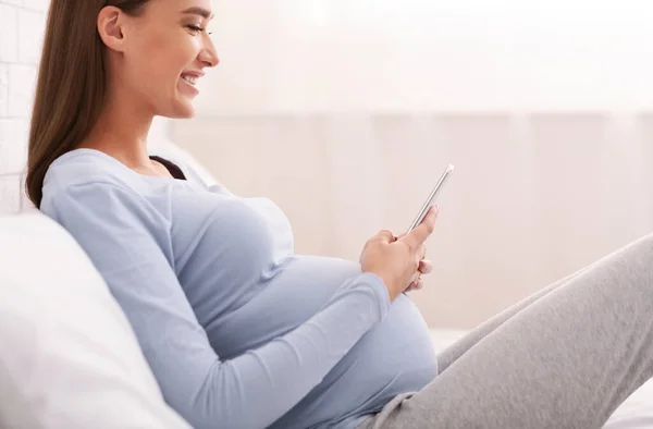 Беременная девушка просматривает на смартфоне, сидя на кровати в спальне, обрезанный — стоковое фото