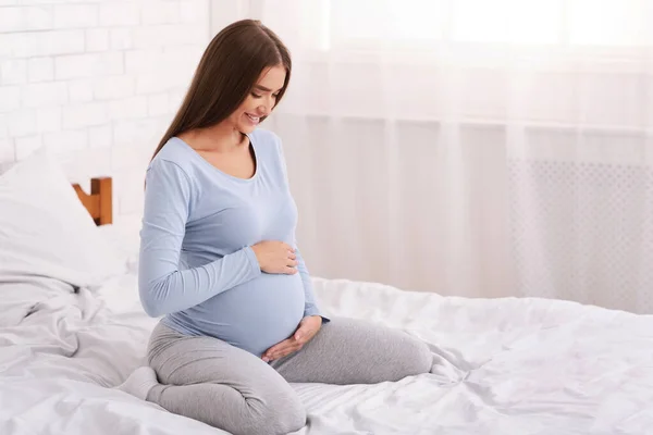 Happy Pregnant Woman Touching Belly Sitting On Bed In Bedroom
