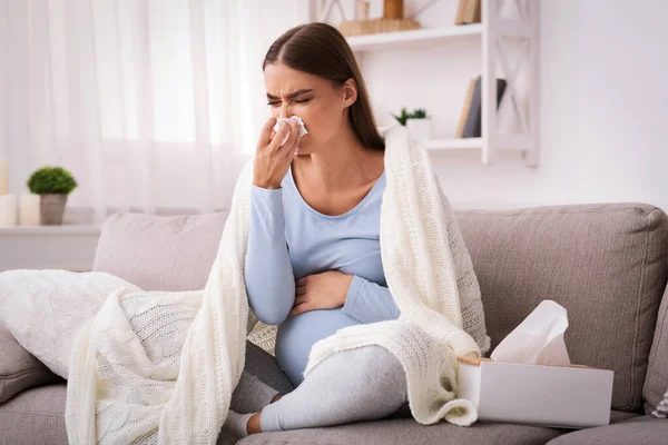 Pregnant woman blowing nose in tissue sitting on sofa indoor — Stock Photo, Image