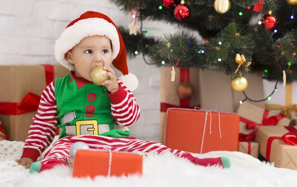 Dítě kousání dekorace míč, sedí pod vánoční strom — Stock fotografie