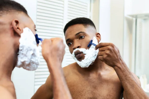 Hombre afroamericano afeitándose en el baño, mirando el espejo — Foto de Stock