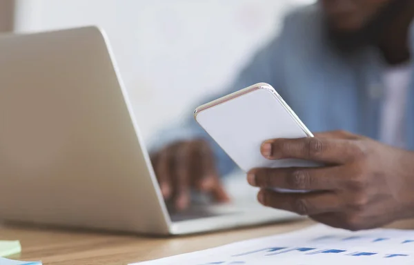 Hombre de negocios afroamericano irreconocible usando teléfono inteligente y escribiendo en el ordenador portátil — Foto de Stock
