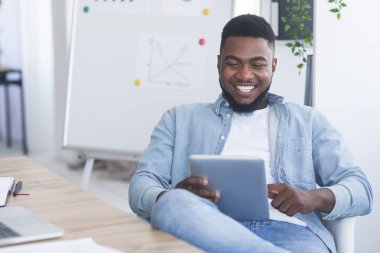 Neşeli Afrikalı iş adamı modern ofiste dijital tabletle oturuyor.
