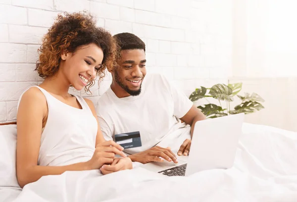 Feliz pareja africana de compras en línea en la cama, utilizando el ordenador portátil y la tarjeta de crédito —  Fotos de Stock