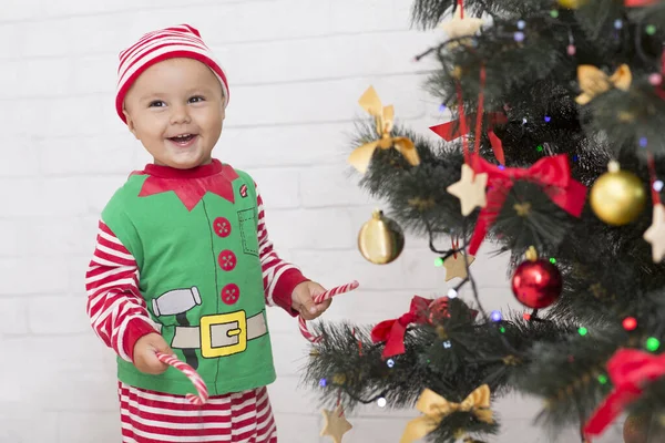 Veselý malý skřítek s lízátkem, smějící se u vánočního stromku — Stock fotografie