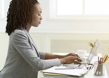 Dizüstü bilgisayarda çalışan genç bayan işyerinde oturuyor, yan görünüm