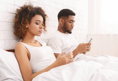 Genç evli Afrikalı çift yatakta cep telefonu kullanıyor.