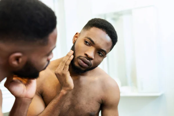 Orolig tusenårig afrikansk kille kollar sitt ansikte, tittar på spegeln — Stockfoto