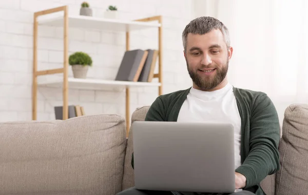 Uomo positivo utilizzando il computer portatile, seduto sul divano a casa — Foto Stock