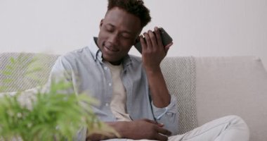 Zenci adam cep telefonundan sesli mesaj dinliyor, evde oturuyor.