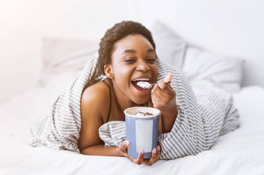 Yatakta dondurma. Gülümseyen Afrikalı Amerikalı kadın battaniyeye sarılıp yemek yiyor.