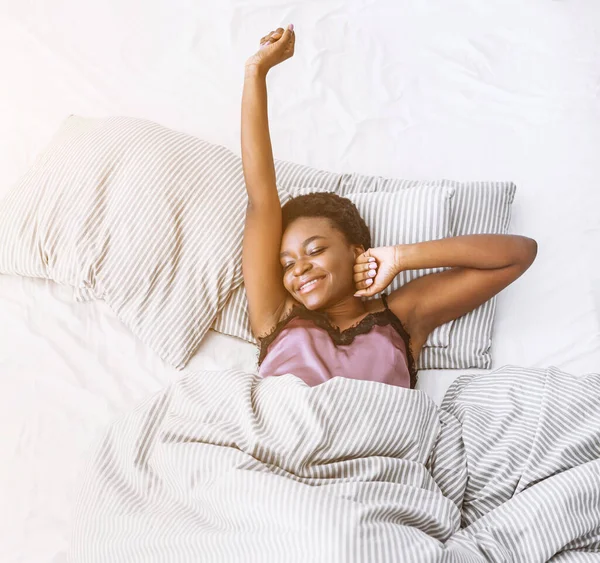 Αρχίζει η ηλιόλουστη μέρα. Χαμογελώντας αφροαμερικανή κοπέλα που τεντώνεται στο κρεβάτι — Φωτογραφία Αρχείου
