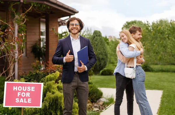 Glücklicher Immobilienmakler zeigt Daumen-hoch-Geste, während sich junges Paar in der Nähe ihres neuen Hauses umarmt — Stockfoto