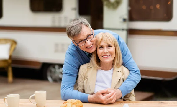 Smiling mature man hugging his beautiful wife in front of camping vehicle outdoors — Stock Photo, Image