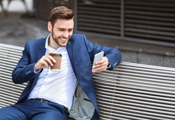 Retrato de jovem empresário usando telefone celular e segurando xícara de café takeout no banco fora, espaço de cópia — Fotografia de Stock