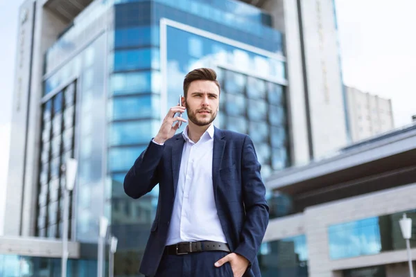 Serious millennial businessman speaking on smartphone near skyscraper at street — Stock Photo, Image