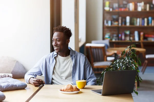 Millennial ragazzo afroamericano con smartphone e laptop godendo mattina caffè e croissant in caffè — Foto Stock