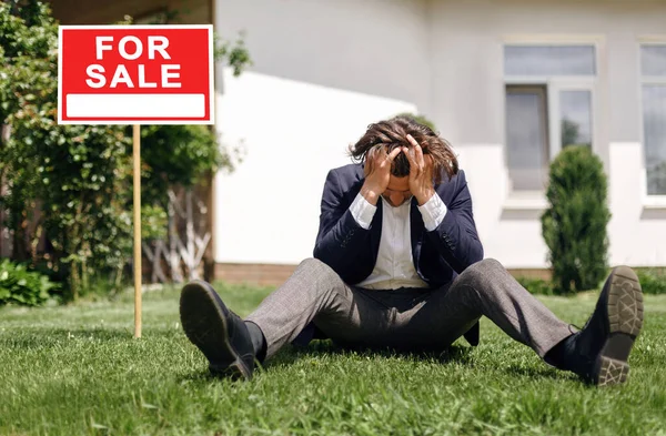 Depressiver Immobilienmakler sitzt auf Gras vor dem Haus zum Verkauf, im Freien — Stockfoto