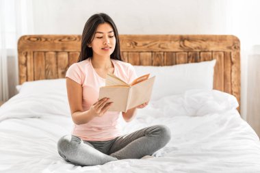 Asyalı kız evde yatağında kitap okuyor.