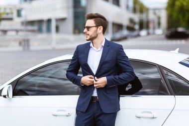 Beyaz takım elbiseli bir işadamı şehir merkezinde yeni arabasının yanında duruyor.