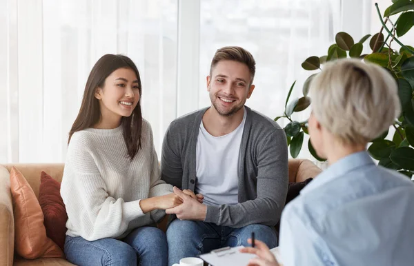 Sonrientes cónyuges multiétnicos sosteniendo las manos reconciliando la sesión en la oficina de psicoterapeutas — Foto de Stock