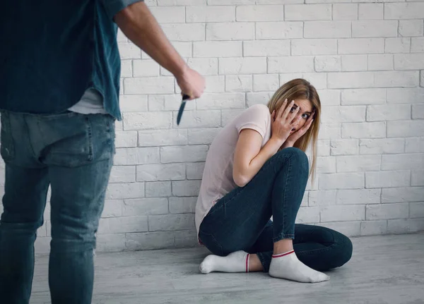 Ameaça de vida em casa. Mulher de horror senta-se no chão, homem segura faca nas mãos — Fotografia de Stock