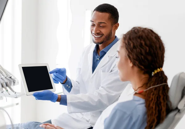 Χαμογελώντας αρσενικό οδοντίατρο δείχνει γυναίκα ασθενή άδειο ψηφιακή οθόνη δισκίο — Φωτογραφία Αρχείου
