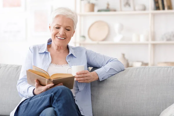 Ideje nyugdíjba menni. Idős hölgy ül a kanapén könyvvel és kávéval — Stock Fotó