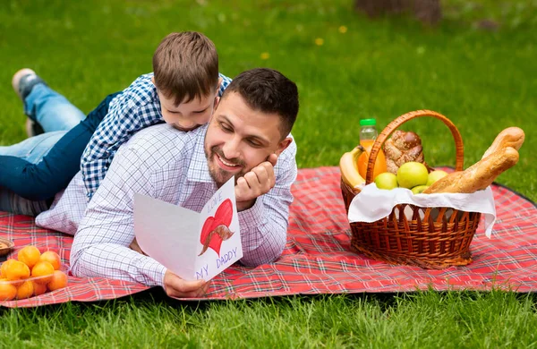 Saludo del día de los mejores padres. Padre e hijo con tarjeta abrazándose a cuadros en el parque durante el picnic — Foto de Stock