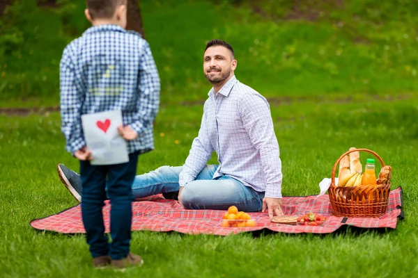 Pequeño niño escondiendo la tarjeta de felicitación hecha a mano detrás de su espalda mientras felicita a su papá con el Día del Padre en el picnic — Foto de Stock