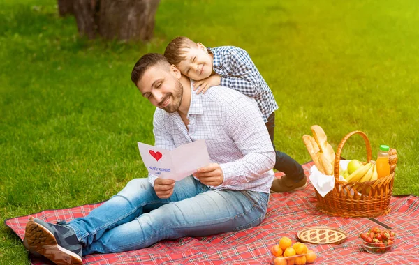 Menino afetuoso abraçando seu pai com cartão de dia de pais artesanais no piquenique ao ar livre — Fotografia de Stock