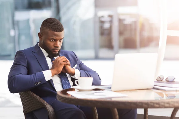 Pensativo hombre de negocios negro sentado en el ordenador portátil pensando en el café al aire libre —  Fotos de Stock