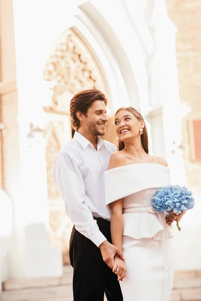 Pareja de boda de pie cerca de la iglesia sosteniendo las manos y abrazando, Vertical — Foto de Stock